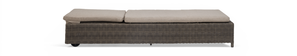Lang rektangulær udendørs skammel/sofa i brun flettet rattan med to beige hynder, frilagt mod sort baggrund.