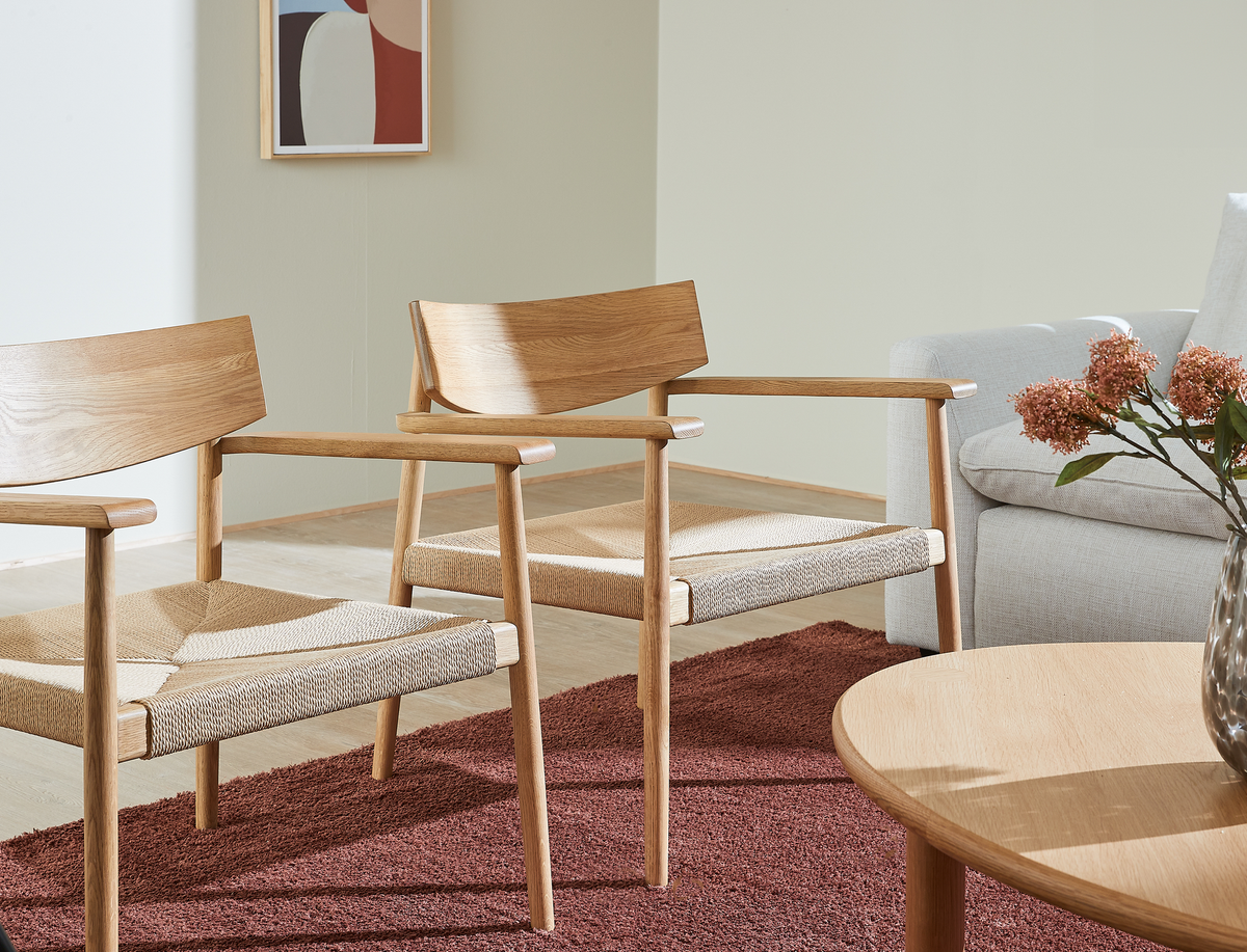 To træstole med flettet sæde på rødbrunt tæppe ved siden af en lysegrå sofa, rund træbordplade og en vase med lyserøde blomster; abstrakt vægbillede på væggen.