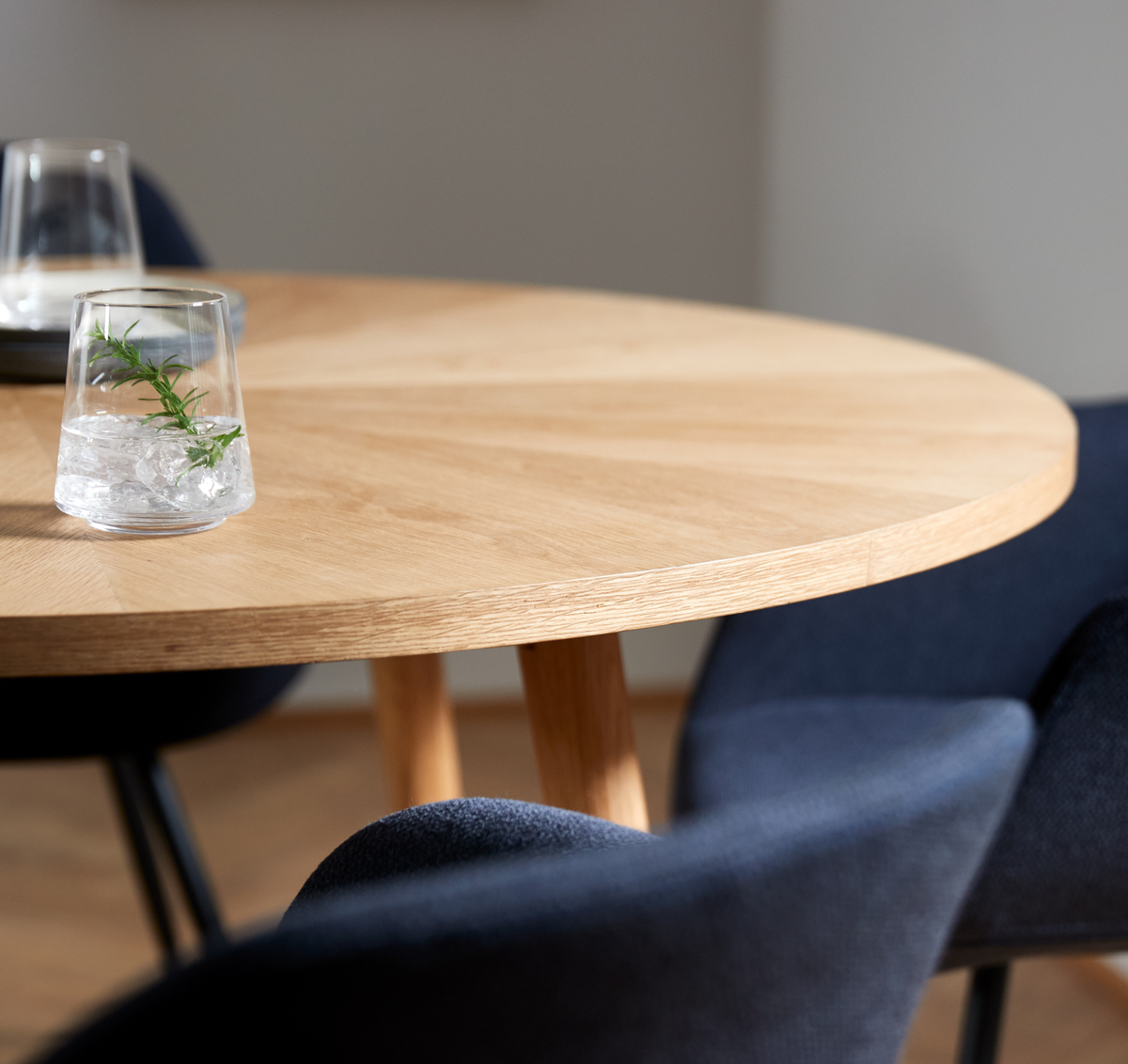 Nærbillede af rund bordplade i lys eg med blå polstrede stole; et glas med is og en rosmarinkvist.