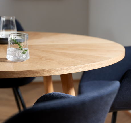 Nærbillede af rund bordplade i lys eg med blå polstrede stole; et glas med is og en rosmarinkvist.