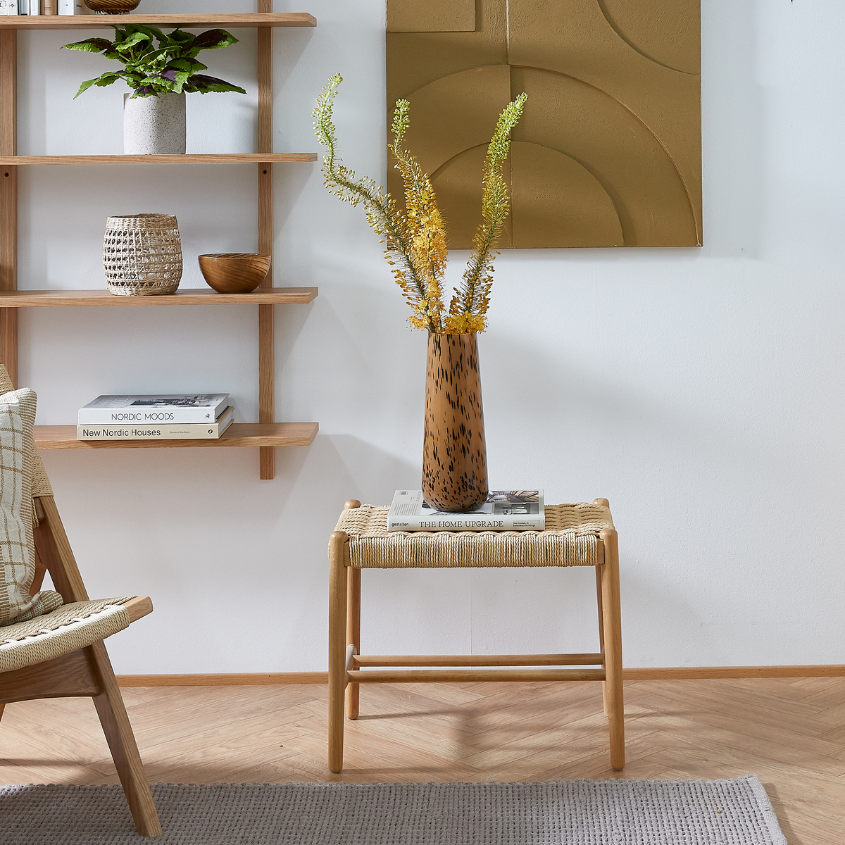 Lyst nordisk stuehjørne: fletskammel med to bøger, ”THE HOME UPGRADE” og et magasin, med brunplettet vase med gule grene; væghylder med plante, kurv og bøger ”NORDIC MOODS” og ”New Nordic Houses” i neutrale træ- og beige toner.