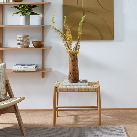 Lyst nordisk stuehjørne: fletskammel med to bøger, ”THE HOME UPGRADE” og et magasin, med brunplettet vase med gule grene; væghylder med plante, kurv og bøger ”NORDIC MOODS” og ”New Nordic Houses” i neutrale træ- og beige toner.