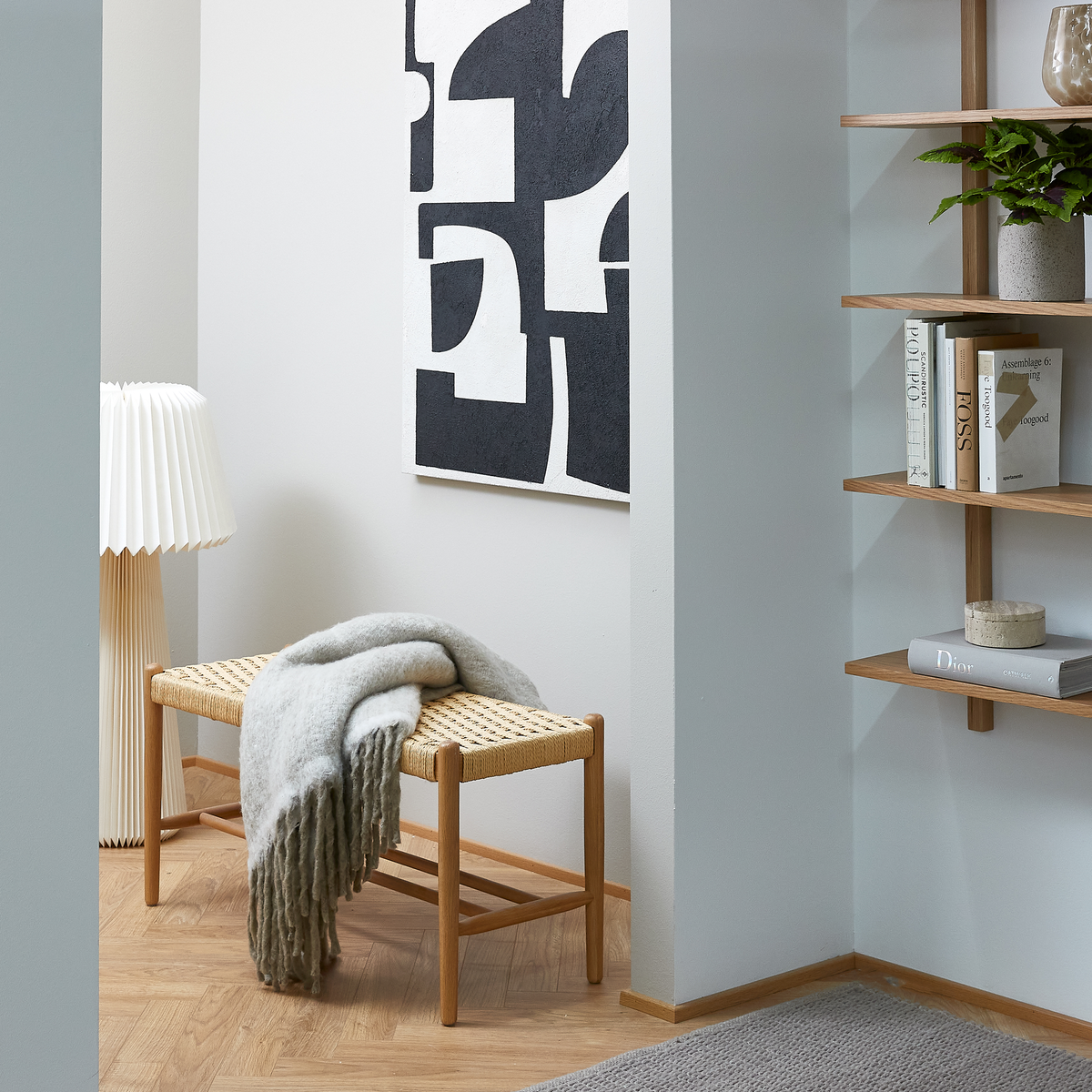Lyst hjørne i entreen med sort-hvid abstrakt kunst, fletbænk med gråt plaid, plisseret gulvlampe og åbne træreoler med bøger som Dior og en grøn plante.