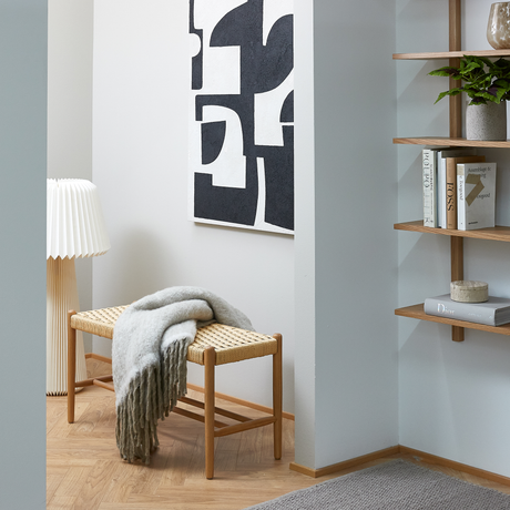 Lyst hjørne i entreen med sort-hvid abstrakt kunst, fletbænk med gråt plaid, plisseret gulvlampe og åbne træreoler med bøger som Dior og en grøn plante.