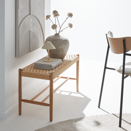 Lys, minimalistisk indretning med flettet bænk, bog og lille bordlampe, stor vase med tørrede blomster og en stol i neutrale nuancer.