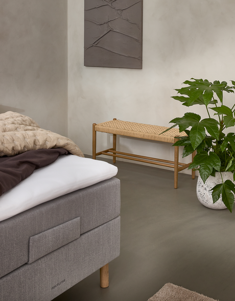 Roligt soveværelseshjørne med grå polstret seng med “nocturne”-mærke, beige plaid, flettet træbænk og grøn potteplante i hvid krukke på neutralt gulv.