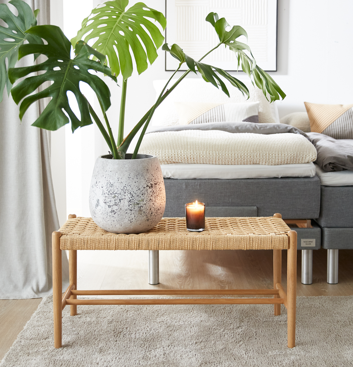 Flettet bænk med monstera i grå krukke og tændt lys foran en grå seng med neutrale tekstiler i et lyst soveværelse.