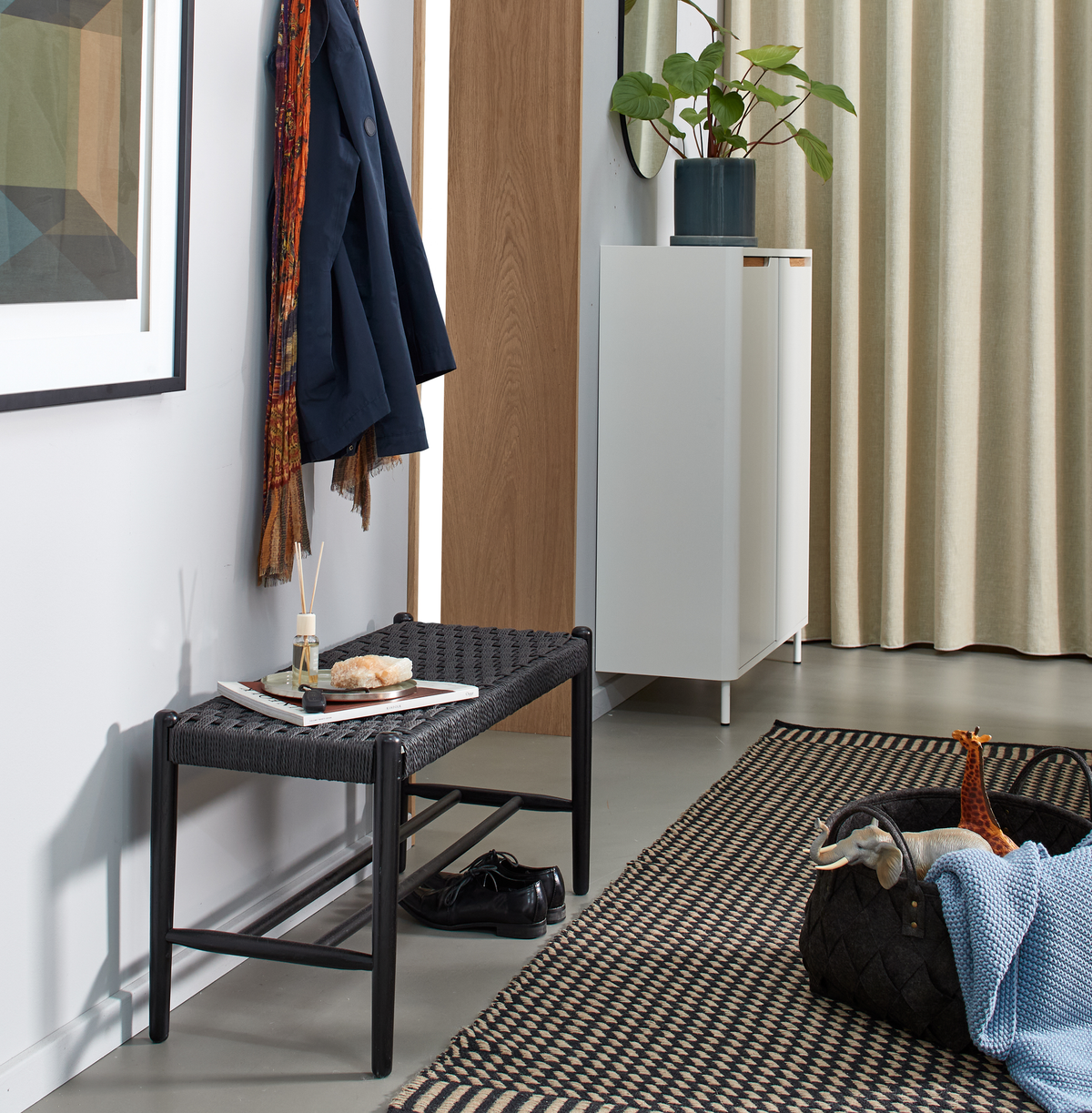Entré med sort flettet bænk, frakke og tørklæde på knage, hvidt skab med grøn plante, mønstret tæppe og kurv med legetøj og plaid; sko under bænken og duftpinde på bakke.