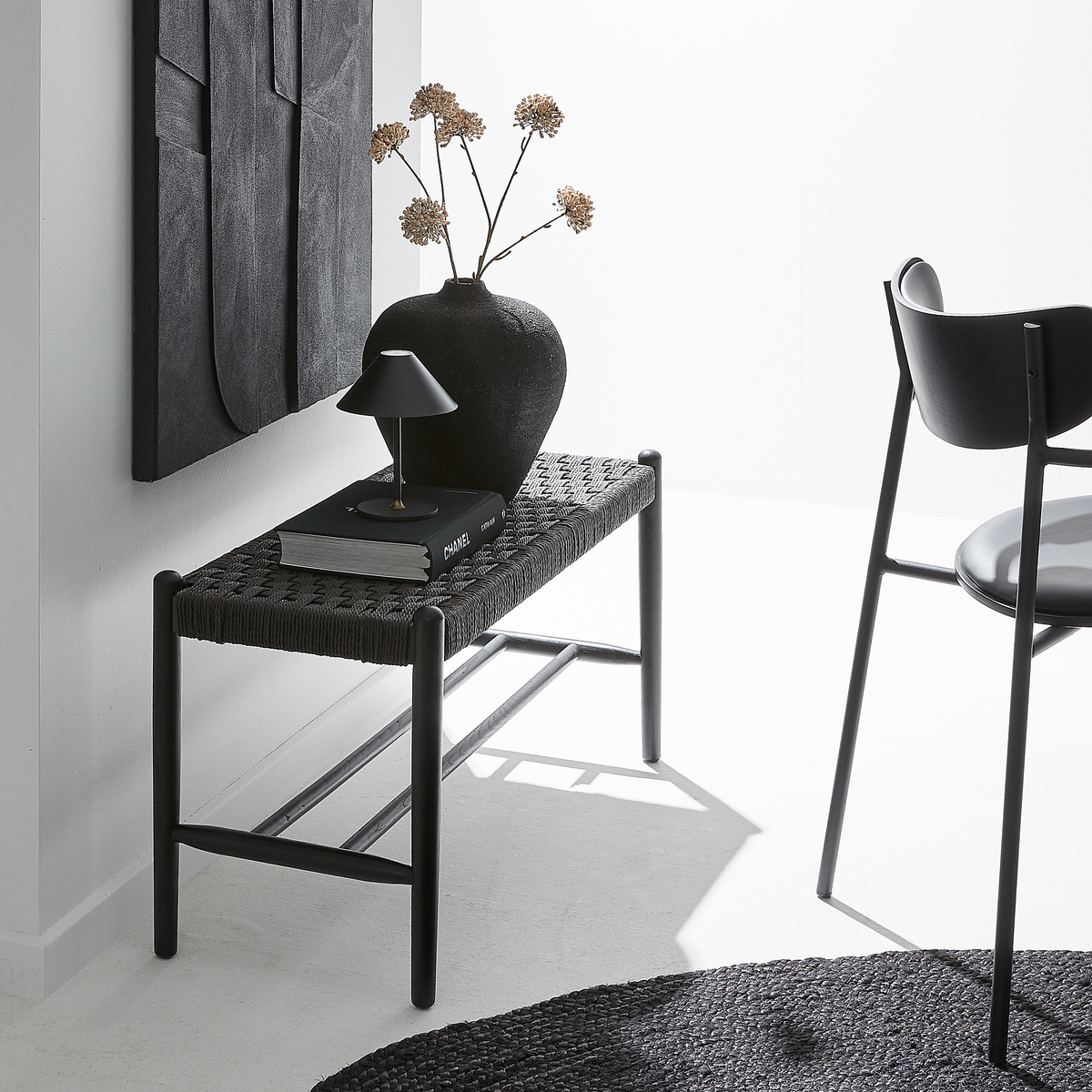 Monokrom hjørne med sort flettet bænk, lille bordlampe, keramikvase med tørrede blomster og en bog med titlen Chanel; sort stol mod hvid væg i stærkt dagslys.