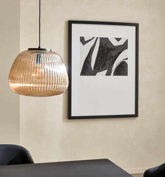 Riflet pendellampe i ravfarvet glas over sort spisebord ved siden af indrammet abstrakt sort-hvid plakat på beige væg.