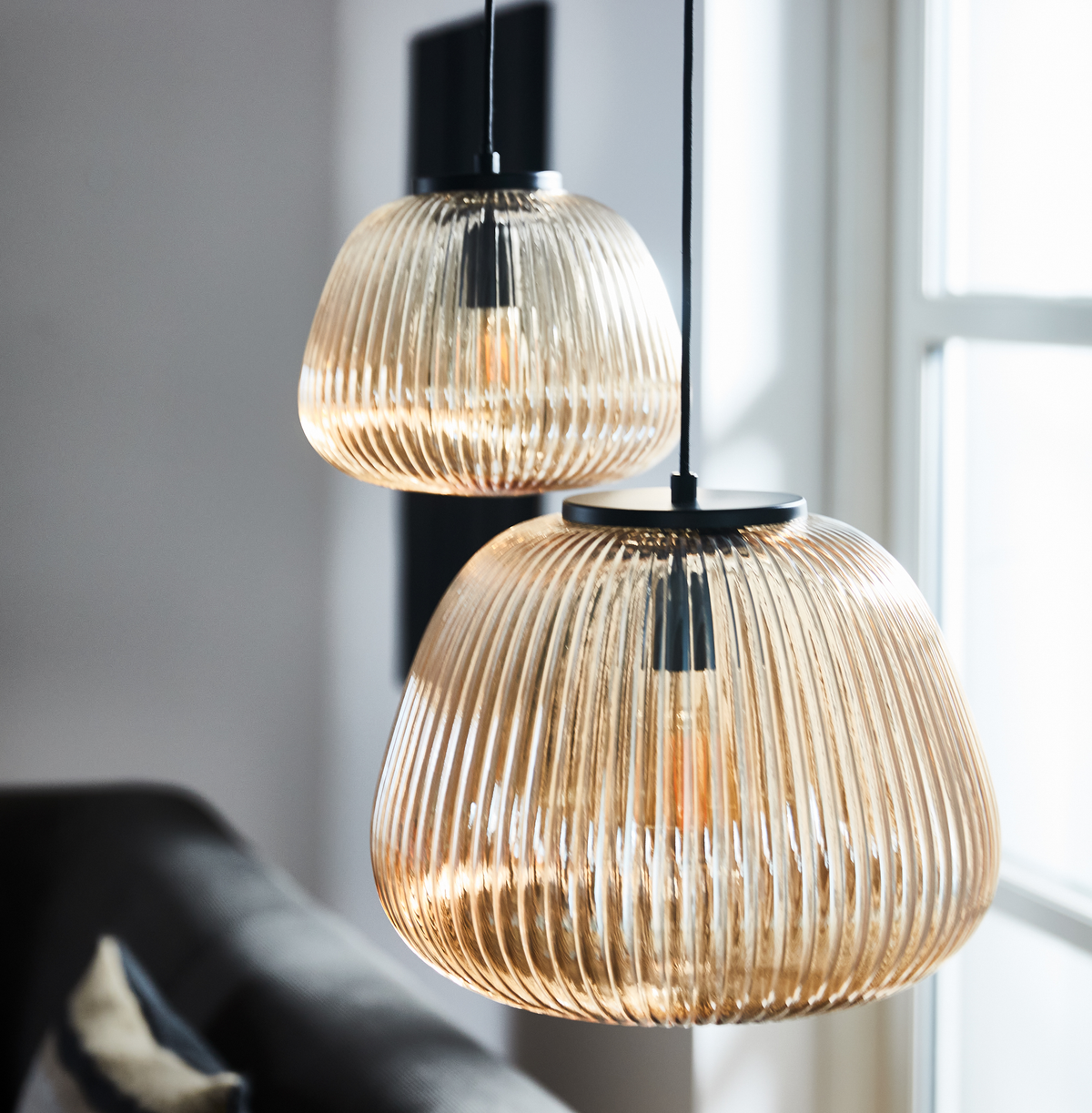 To rillet pendellamper i ravfarvet glas lyser i en moderne stue ved et vindue.