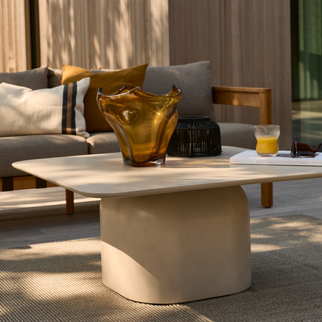 Beige udendørs sofabord med afrundede kanter med en ravfarvet glasvase, sort lanterne, en bog og et glas appelsinjuice; sofa med puder i baggrunden på en terrasse i blødt dagslys.