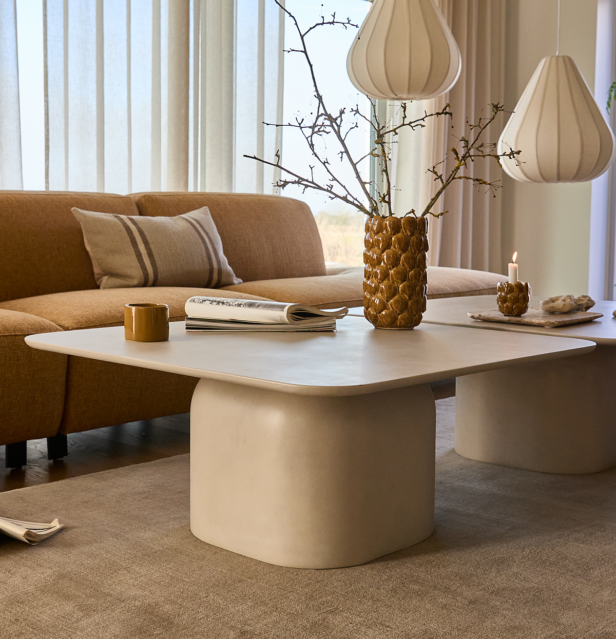 Lyst stue med beige sofabord med afrundede hjørner, sennepsgul sofa, hvide pendellamper, keramikvase med grene, tændt lys, kop og magasin på bordet.