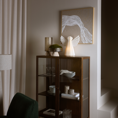Mørkt vitrineskab med glasdøre i en stue; over hænger abstrakt vægkunst, og på skabet står en foldet papirengel-lampe samt en rillet vase med grøn mos; i skabet ses glas, skåle, bøger og to lys, ved siden af lyse gardiner.