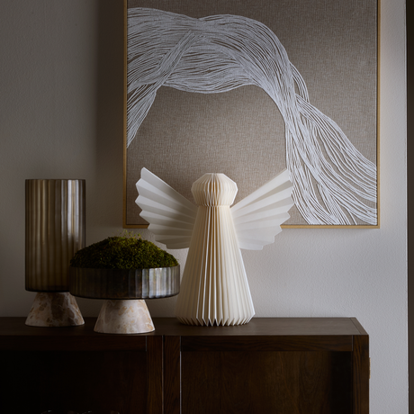 Neutralt stilleben med en foldet papirengel på en mørk trækommode, ved siden af en rillet vase og en lav skål med mos; abstrakt linjekunst i beige og hvid i baggrunden.