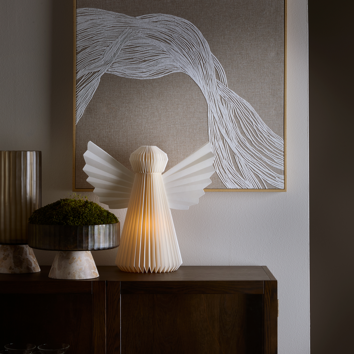 Bordlampe formet som en hvid papirengel lyser på en mørk trækommode foran abstrakt vægkunst, med en dekorativ skål med mos og en riflet vase ved siden af.