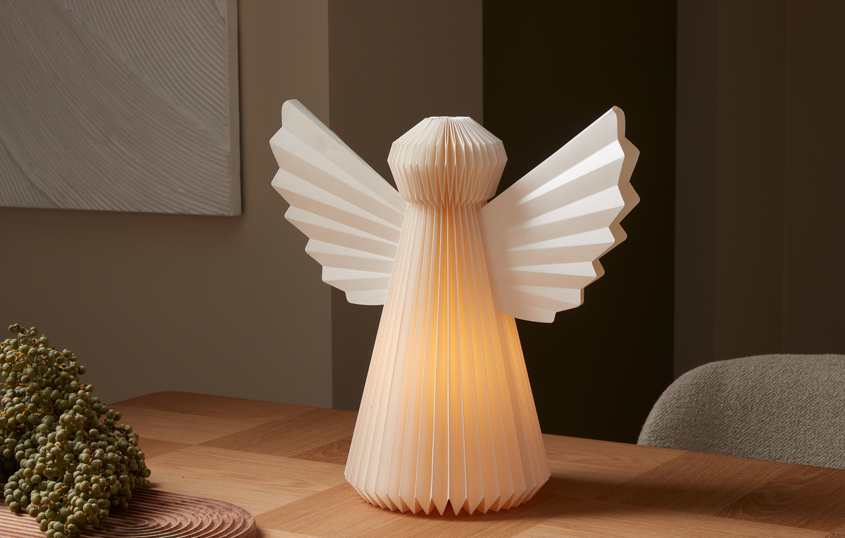 Foldet paplampe formet som en engel med varmt skær på et træbord i et hyggeligt rum.