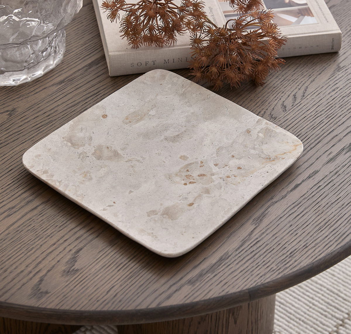 Firkantet beige stenbakke på et mørkt rundt træbord ved siden af en klar glasskål, bogen Soft Minimal og tørrede kviste.