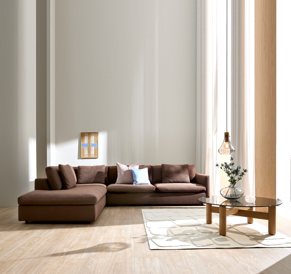 Lyst, minimalistisk stue med brun modulsofa, lyse puder, glas sofabord med træben, vase med blomster og en pendellampe ved store vinduer med hvide gardiner.