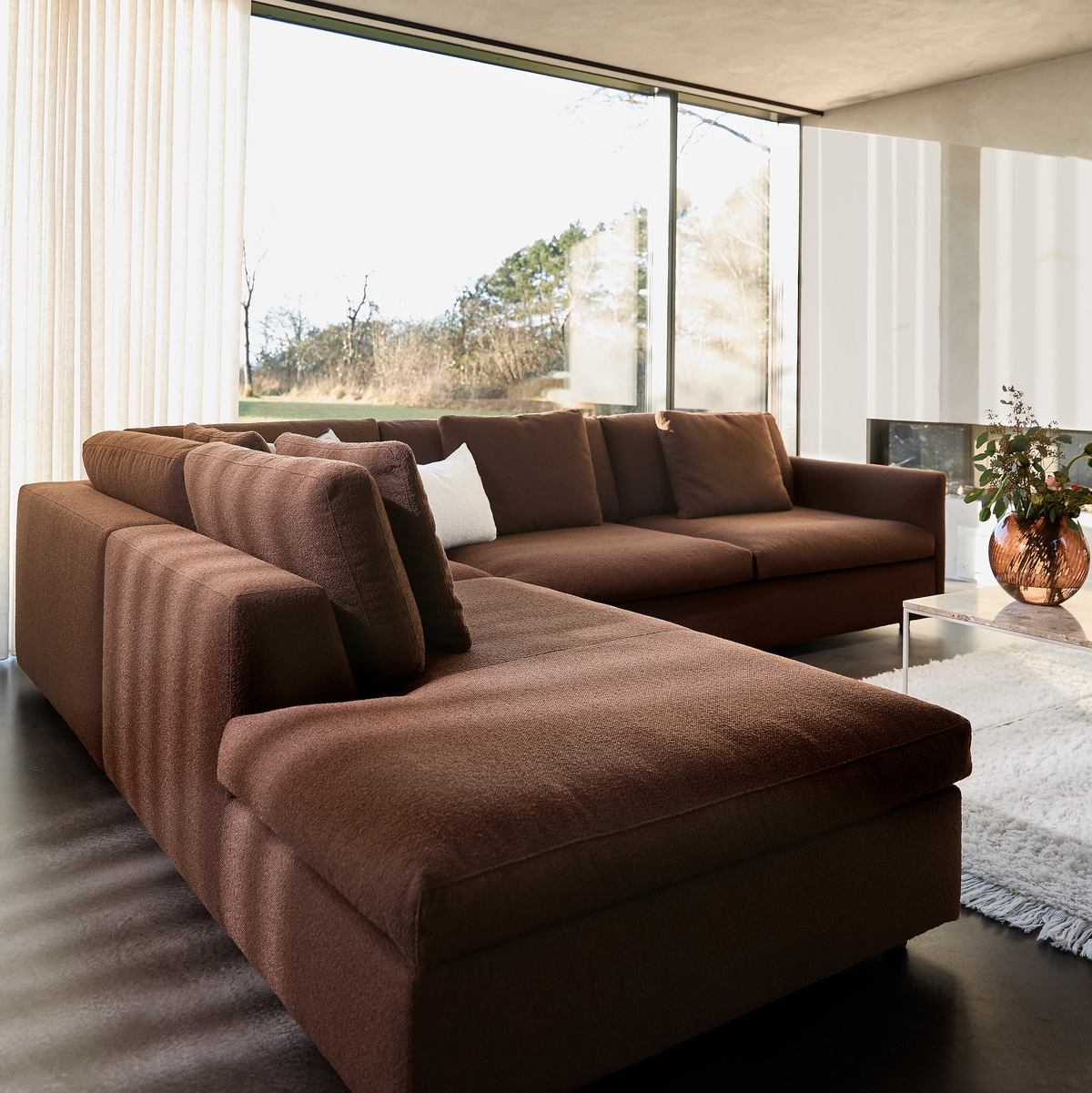 Brun modulsofa i et lyst stuemiljø med gulv‑til‑loft vinduer, hvid rygtæppe og glasbord med kobberfarvet vase og grønne grene.