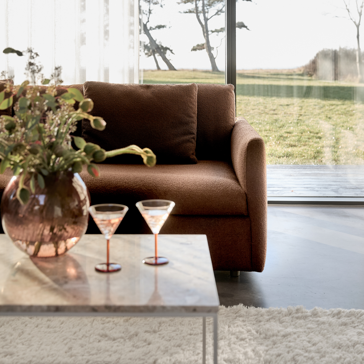 Brun sofa ved gulv-til-loft vindue, marmorbord med glasvase og to cocktailglas, lyst opholdsrum.