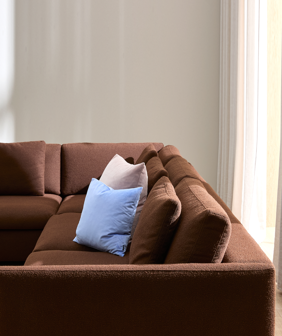 Brun modulsofa med lyseblå og lyserosa pyntepuder mod lys væg og hvide gardiner i dagslys.