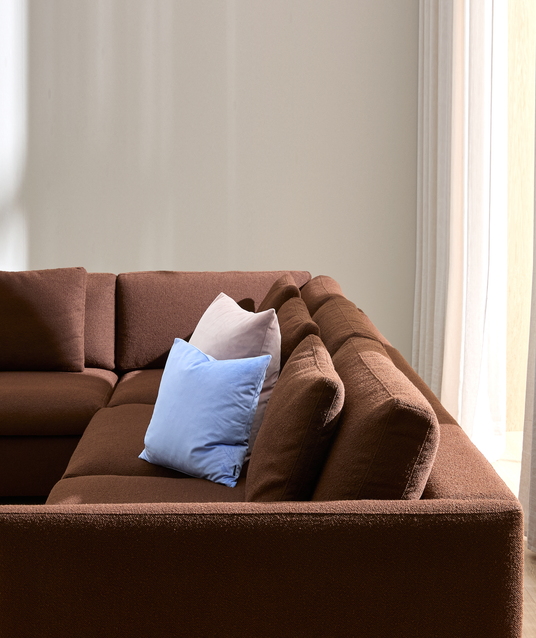 Brun modulsofa med lyseblå og lyserosa pyntepuder mod lys væg og hvide gardiner i dagslys.