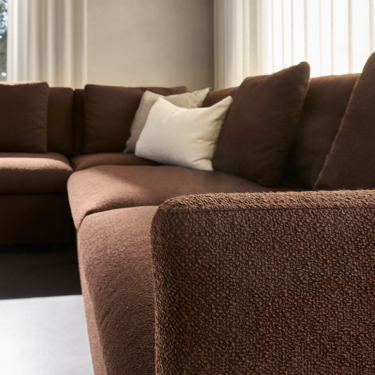 Brun hjørnesofa i bouclé-lignende stof med lys pyntepude i en lys stue med gardiner.