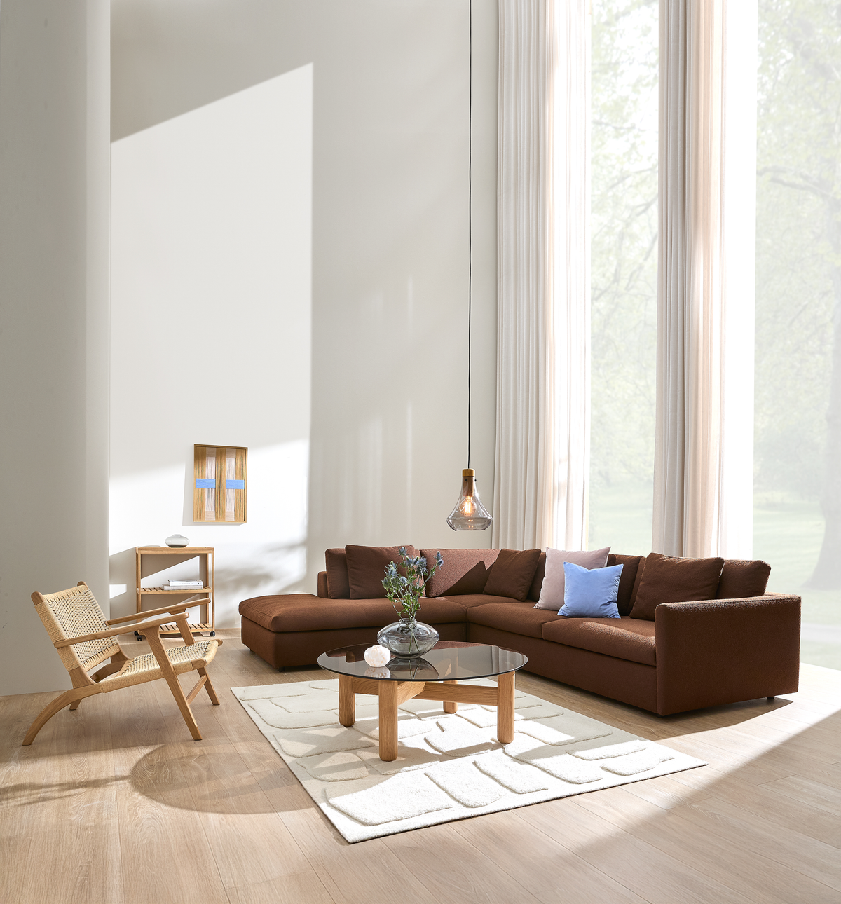 Lyst stue med brun modulsofa, blå accentpude, rundt sofabord med sort glas og træben samt vase med blomster, høje vinduer med lyse gardiner og hængende glaspendel; lyst tæppe og trægulv.