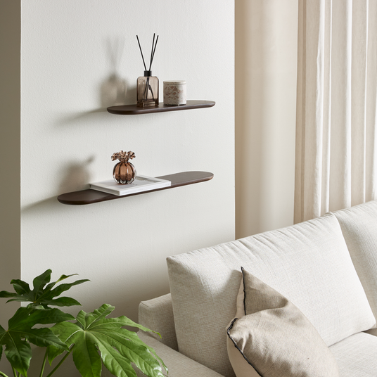Beige sofa mod lys væg med to mørkebrune væghylder med duftpinde, lys og lille vase; grøn plante og lyse gardiner i en lys stue.