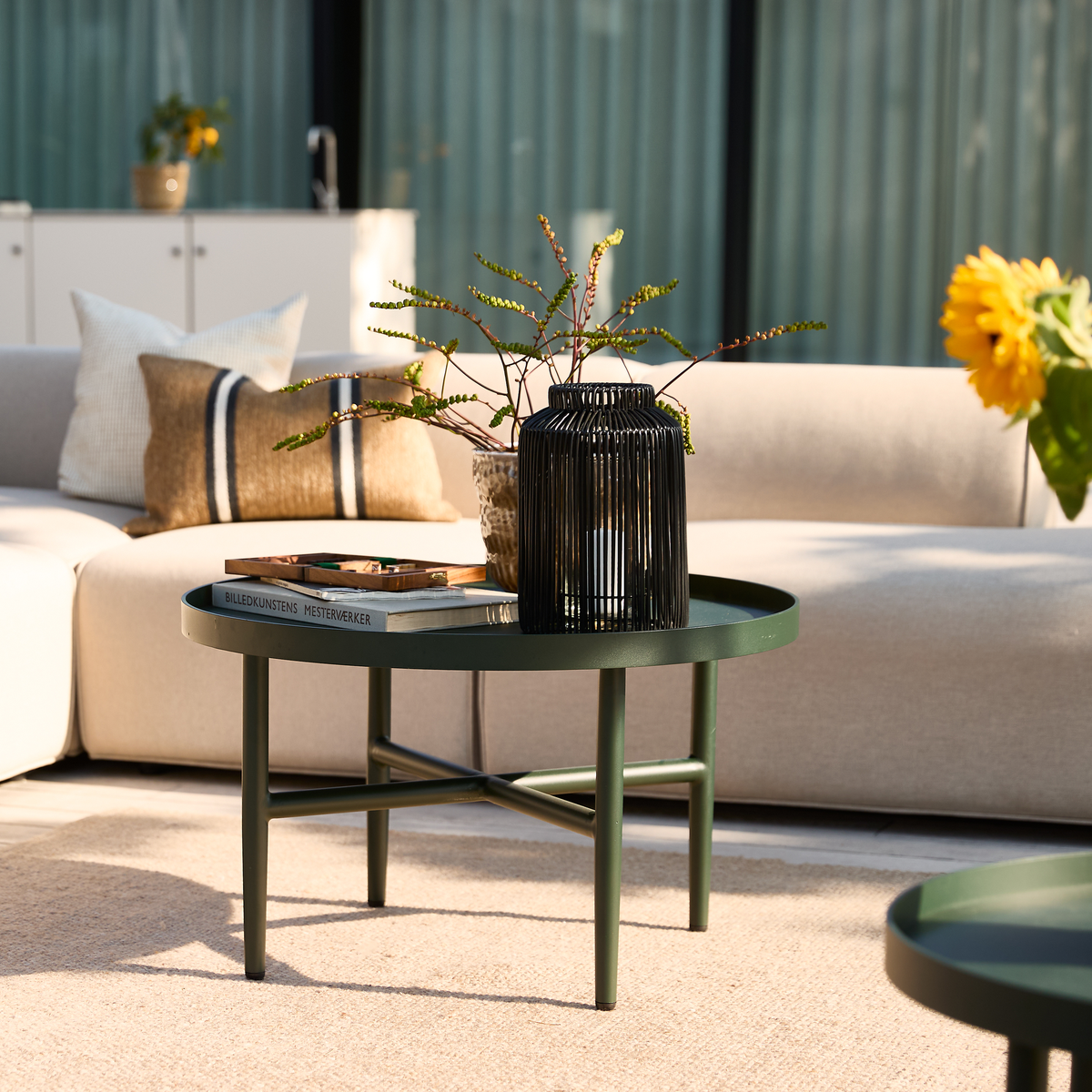 Solbelyst stue med beige sofa; grønt rundt sofabord med sort lanterne, vase med grene og en bog med titlen BILLEDKUNSTENS MESTERVÆRKER.