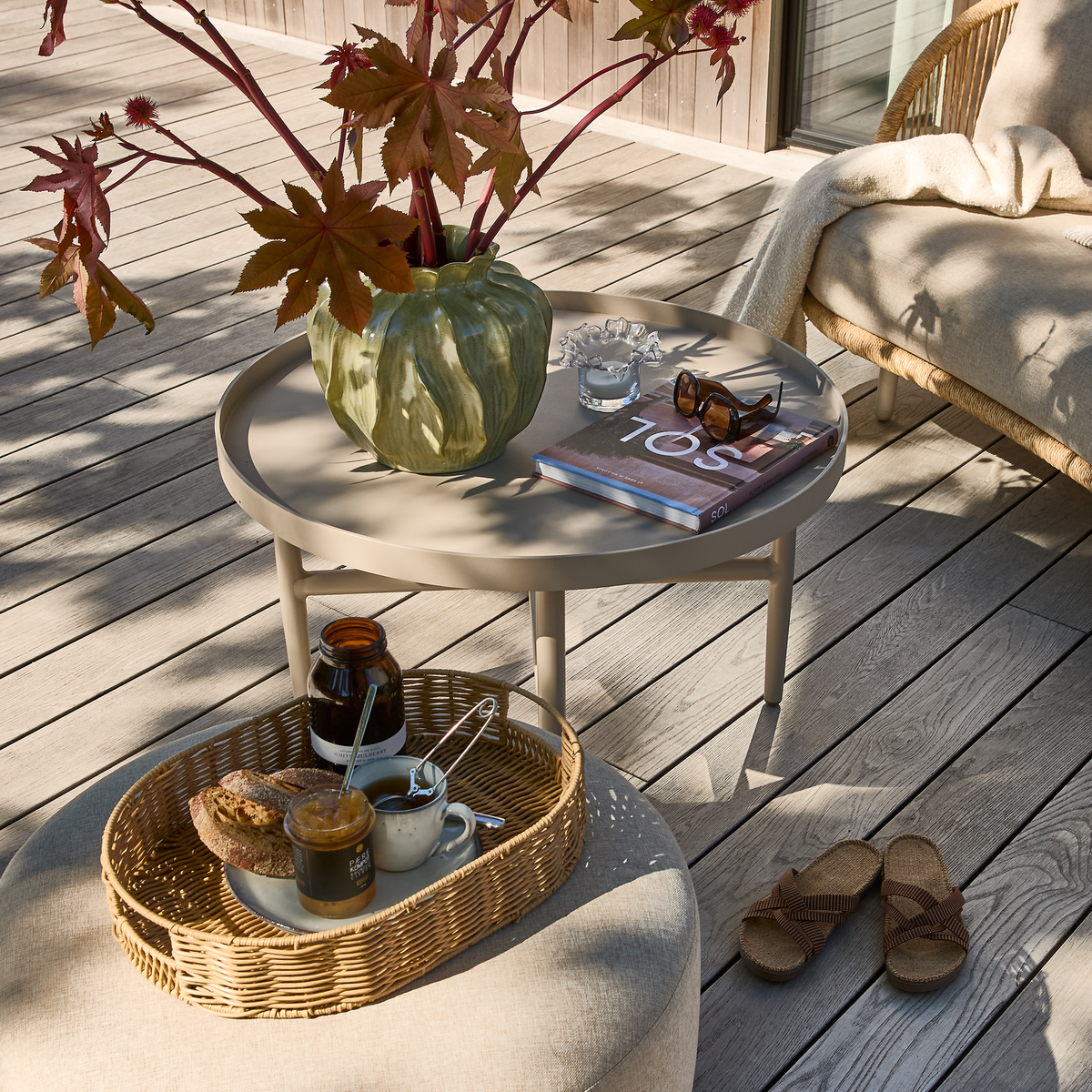 Beige rundt bord på terrasse med grøn vase med røde blade, solbriller og bogen ”TOS”; flettebakke med morgenmad og sandaler ved en loungestol med plaid i sollys.