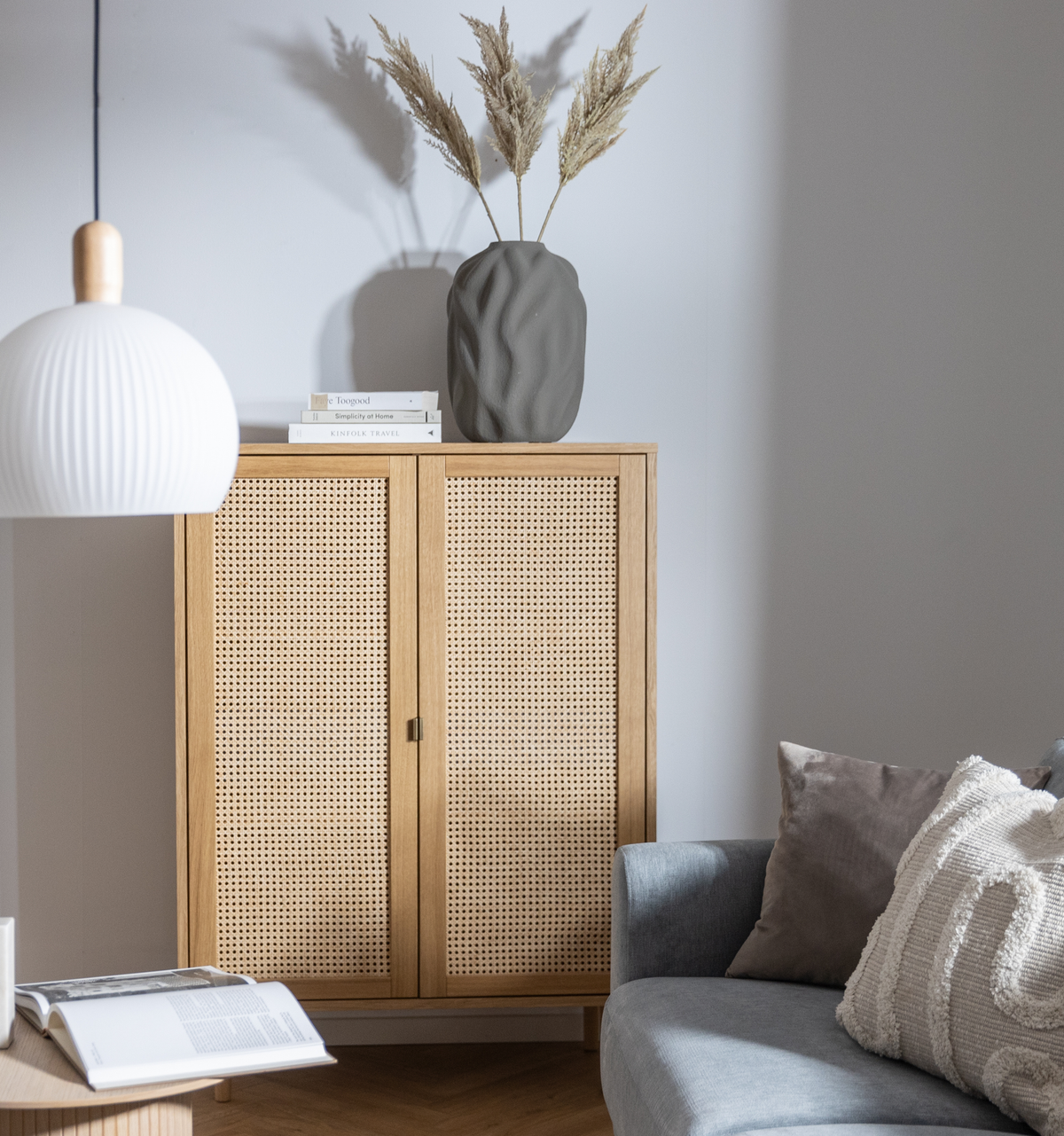 Skandinavisk stuehjørne med egetræsskab med fletfronter, grå sofa med puder, rillet hvid pendel, grå vase med pampasgræs og bøger – bl.a. Kinfolk Travel – ovenpå skabet.
