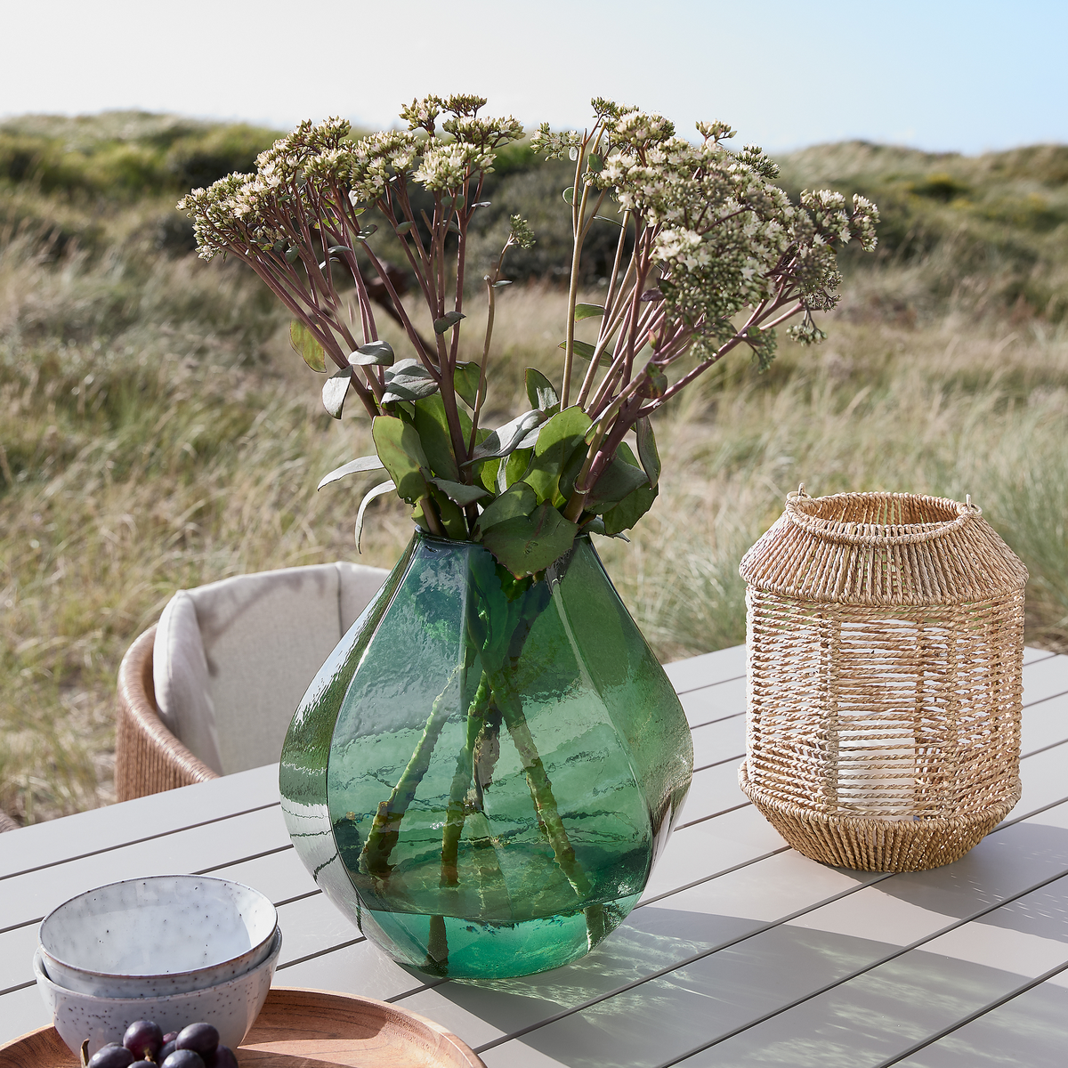 Grøn glasvase med blomster på et udendørs bord ved siden af en flettet lanterne og en skål med druer i dagslys.