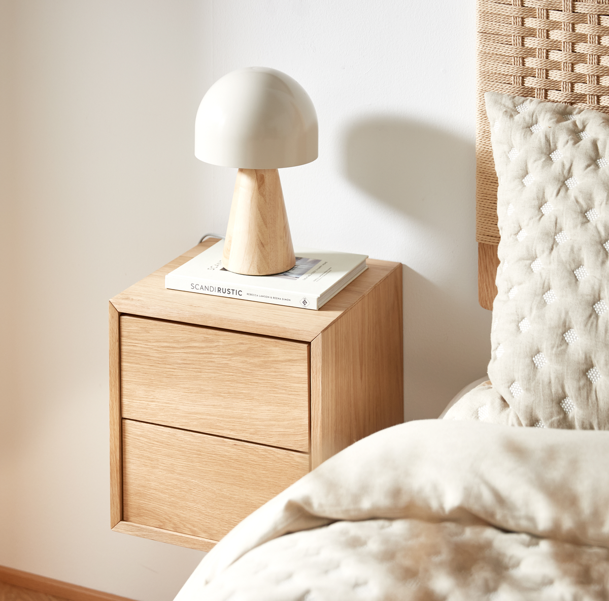 Egetræ natbord med lille bordlampe, en bog med titlen SCANDI RUSTIC og beige sengetøj i et lyst soveværelse.