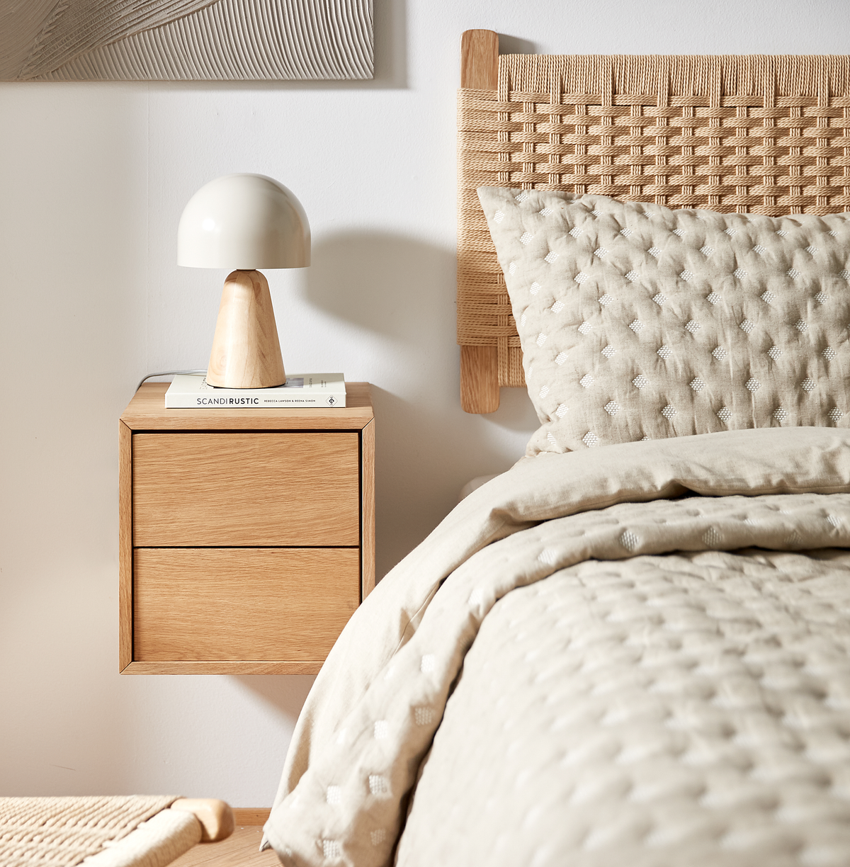 Lyst soveværelsesnærbillede med flettet sengegærde, quiltet beige sengetøj, svævende natbord i eg med kuppelformet bordlampe og bogen SCANDI RUSTIC.