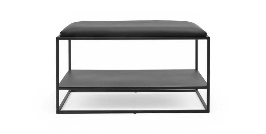 Sort minimalistisk bænk med polstret sæde og åben metalramme med hylde under, frilagt på hvid baggrund.