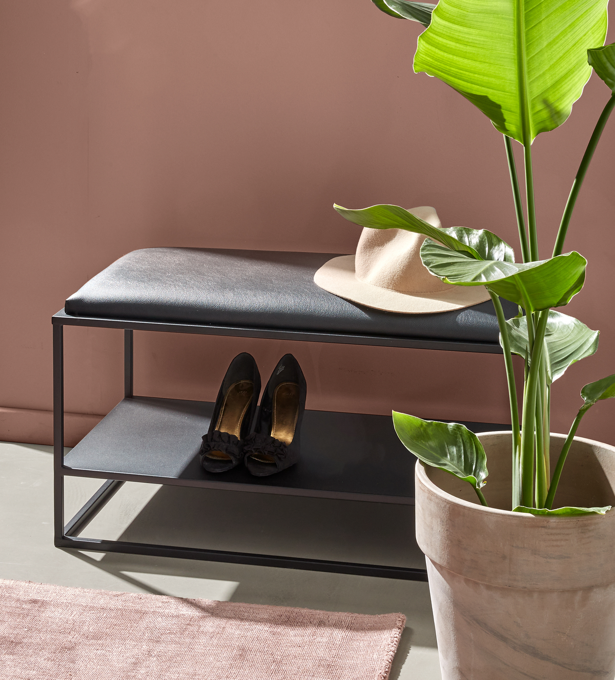 Sort metalbænk med polstret sæde og hylde, en beige hat ligger ovenpå, sorte højhæle på hylden og en grøn potteplante ved siden af mod en rosa væg.