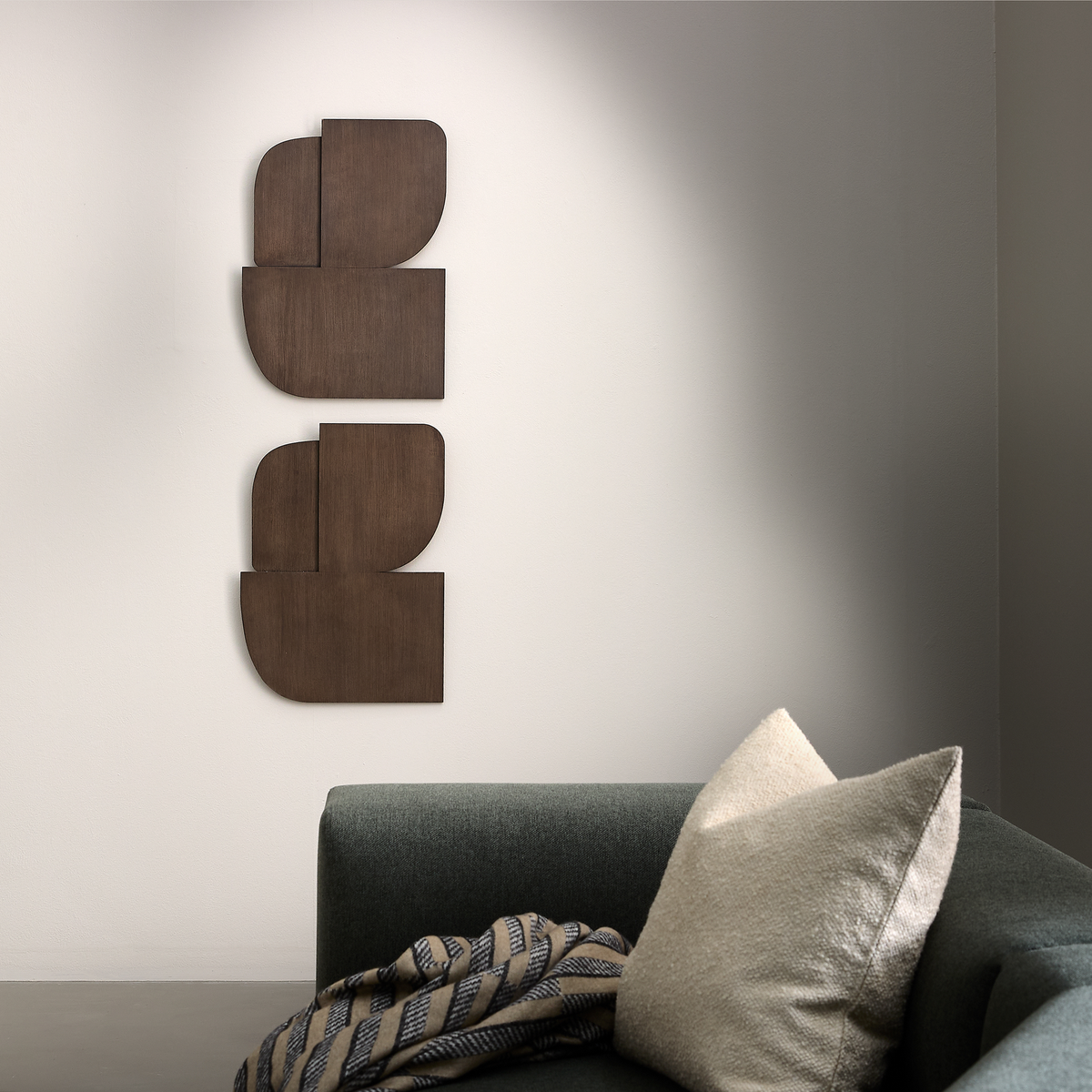 Minimalistisk stue med mørkegrøn sofa, beige pude og grafisk vægkunst i mørkt træ på en hvid væg.