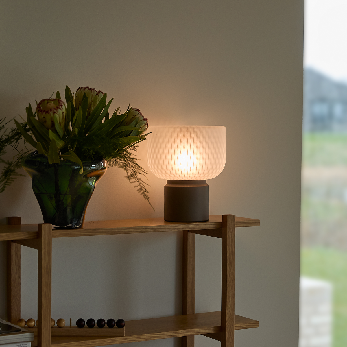 Tændt bordlampe i frostet glas på en træreol ved siden af en grøn vase med blomster ved et vindue.