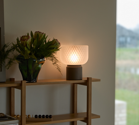 Bordlampe med blødt skær ved siden af grøn glasvase med blomster på træreol i stuen med dagslys fra vindue.
