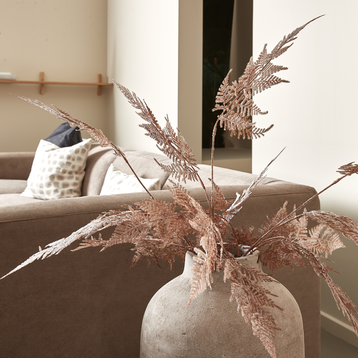 Stor betonvase med tørrede bronzefarvede bregnegrene ved siden af en beige sofa med puder i en lys stue.