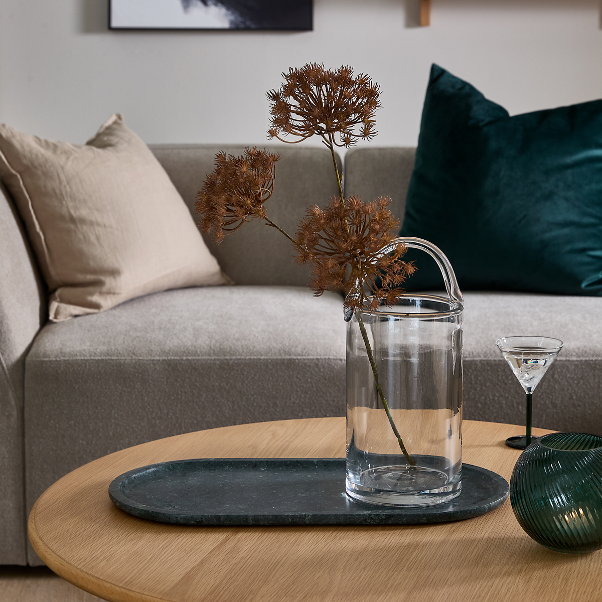 Glasvase med tørrede grene på mørk oval bakke på rundt sofabord i træ; beige sofa med puder og grøn velourpude i baggrunden samt lille cocktailglas og grøn riflet dekor.