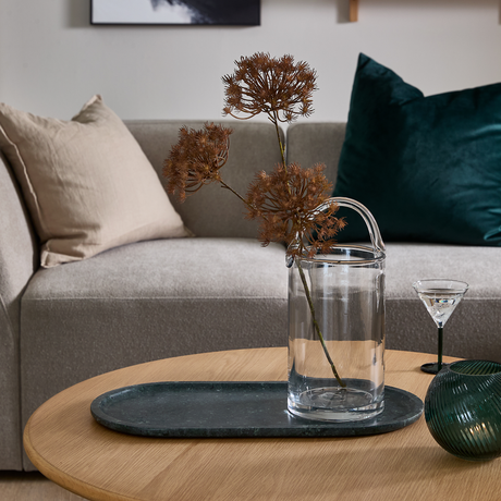 Glasvase med tørrede grene på mørk oval bakke på rundt sofabord i træ; beige sofa med puder og grøn velourpude i baggrunden samt lille cocktailglas og grøn riflet dekor.