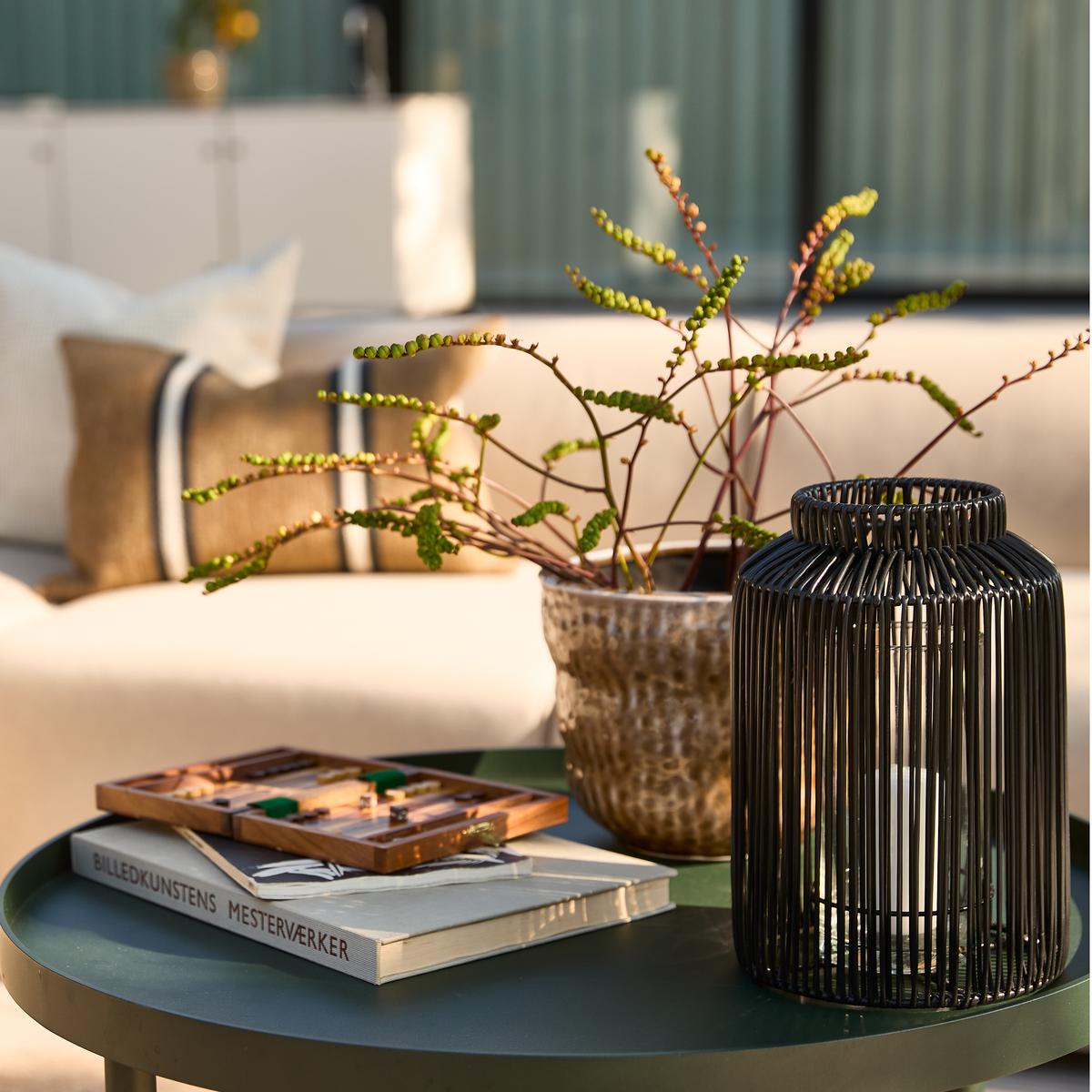 Rundt grønt bakkebord med sort lanterne, keramikvase med grønne grene, et træspil og en bog med titlen ”Billedkunstens Mesterværker”, med lys sofa i baggrunden i varmt dagslys.