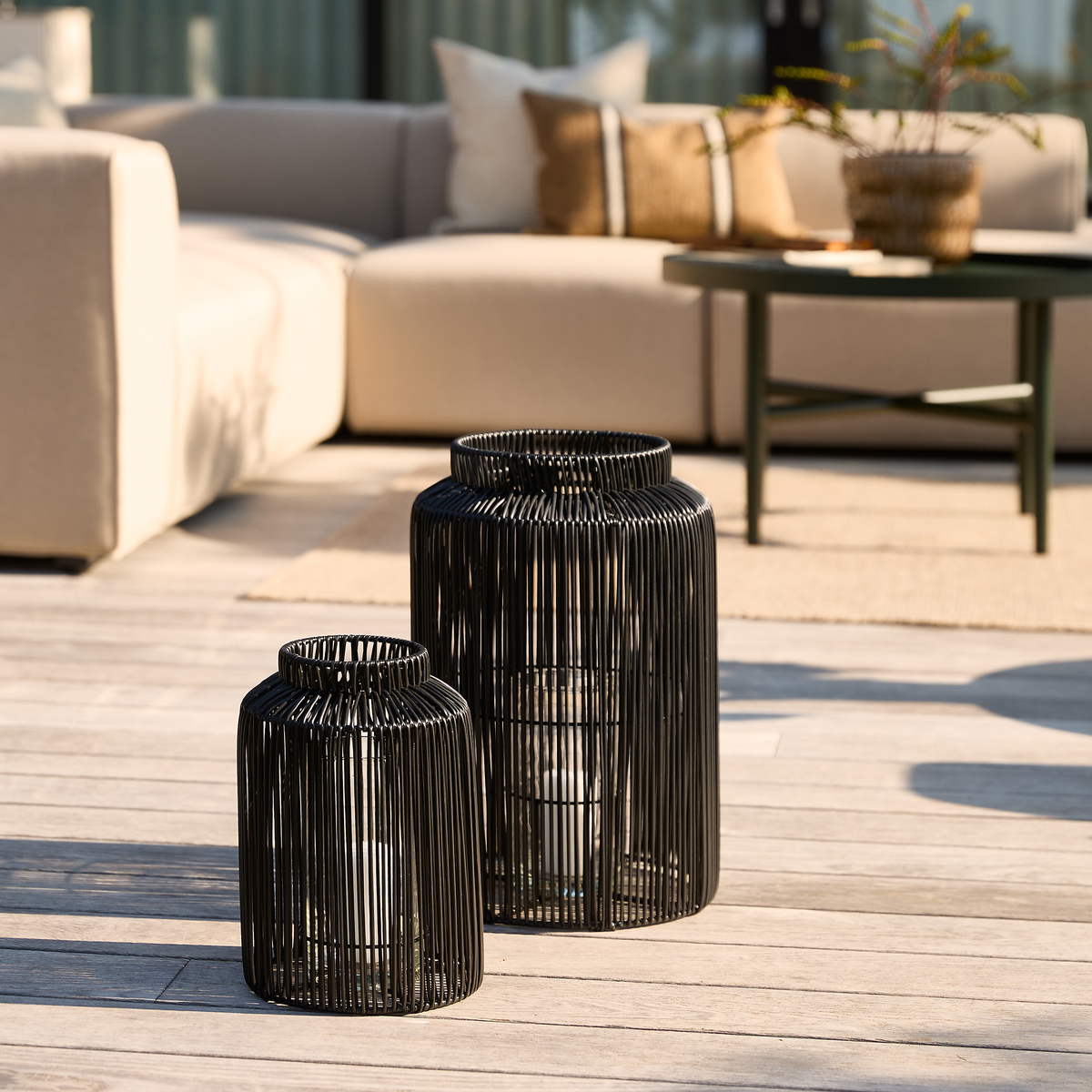 To sorte metallanterner med stearinlys på en solrig terrasse foran en beige loungesofa og grønt sofabord.