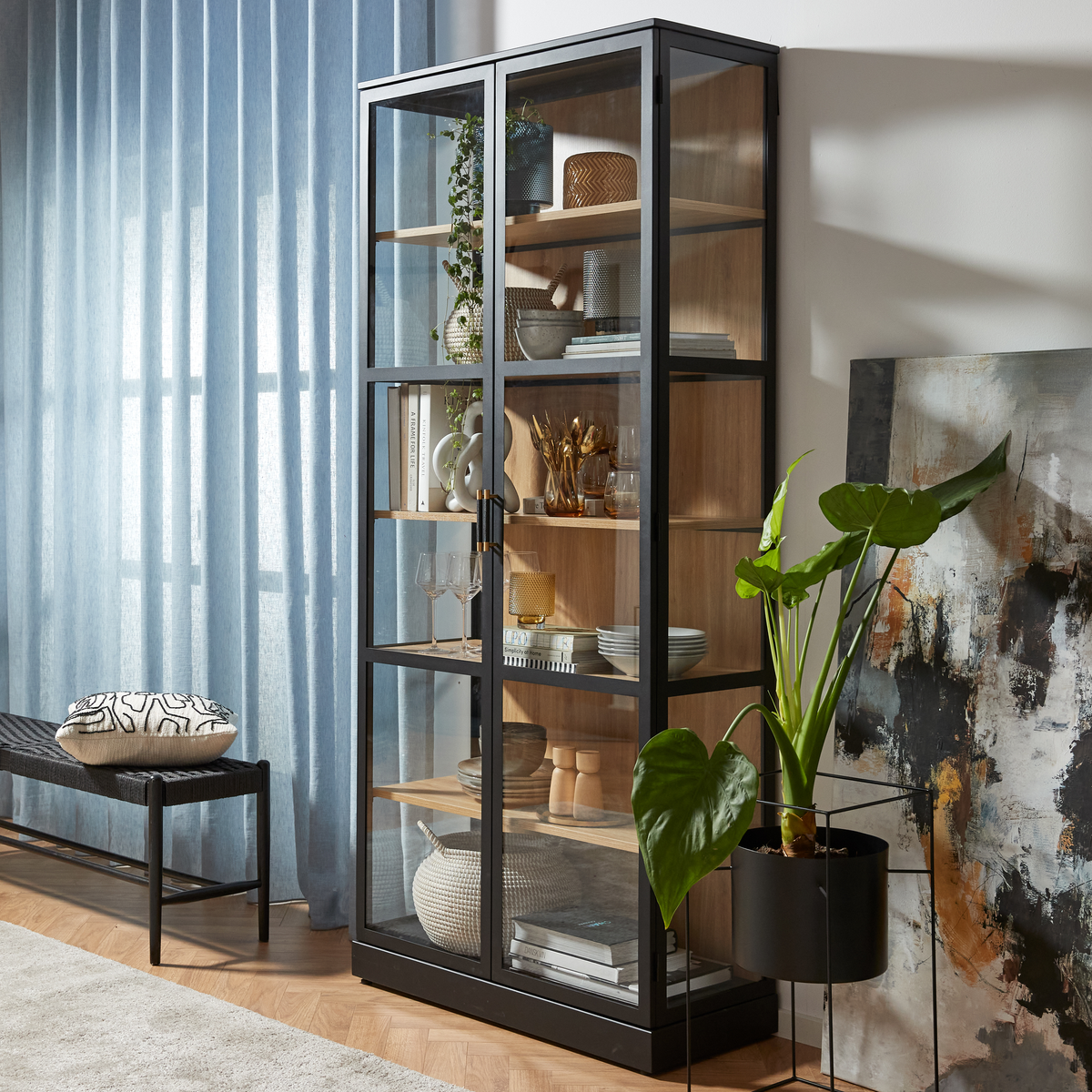 Sort vitrineskab med glaslåger og træhylder, stylet med keramik, glas, bøger og skåle; stor grøn potteplante, lyseblå gardiner og et abstrakt maleri i en moderne stue.