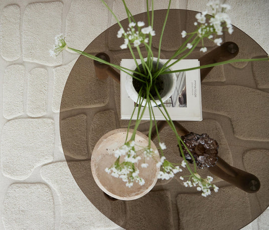 Set oppefra af et rundt glas-sofabord med hvide blomster i vase, en bog, en lys stenskål og et brunt glasobjekt på et beige, struktureret tæppe.