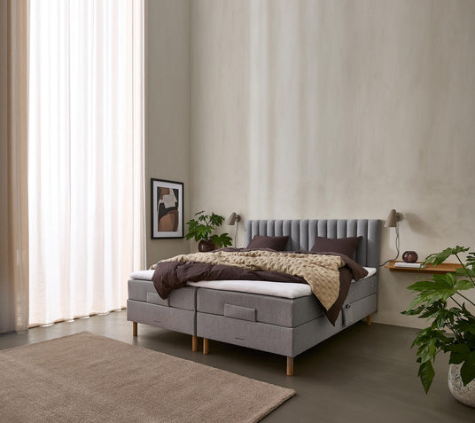 Lyst soveværelse med grå polstret seng, beige quilt og mørkebrunt sengetøj, vægmonterede lamper, planter og lyse, lette gardiner.