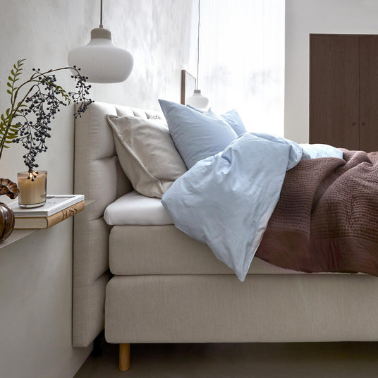 Lyst soveværelse med beige polstret seng, lyseblåt sengetøj, brun strikket plaid, pendel, gren i vase, tændt duftlys og bogen PIGS på hylden.